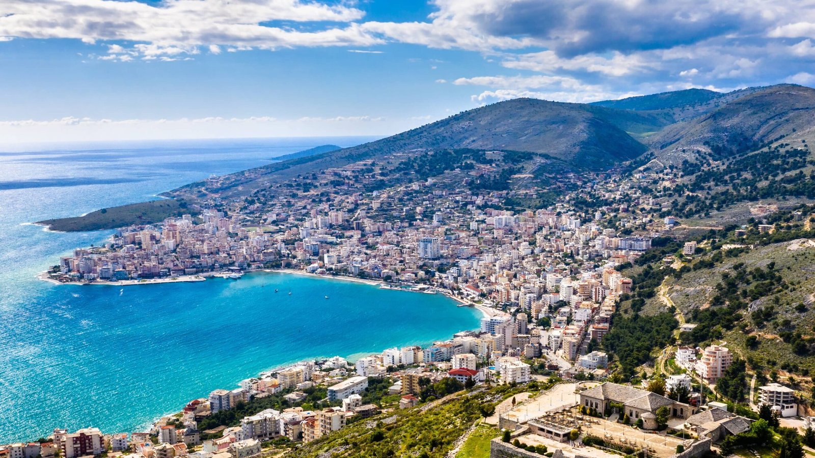 aerial view of Saranda Bay in Albania, featuring the stunning blue-green waters of the Ionian Sea, buildings, harbour and mountains 