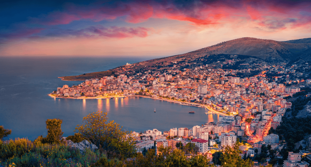 Sunset panorama over Saranda, capturing the bay, harbor, cityscape, promenade, architectural highlights, and the shimmering Ionian Sea