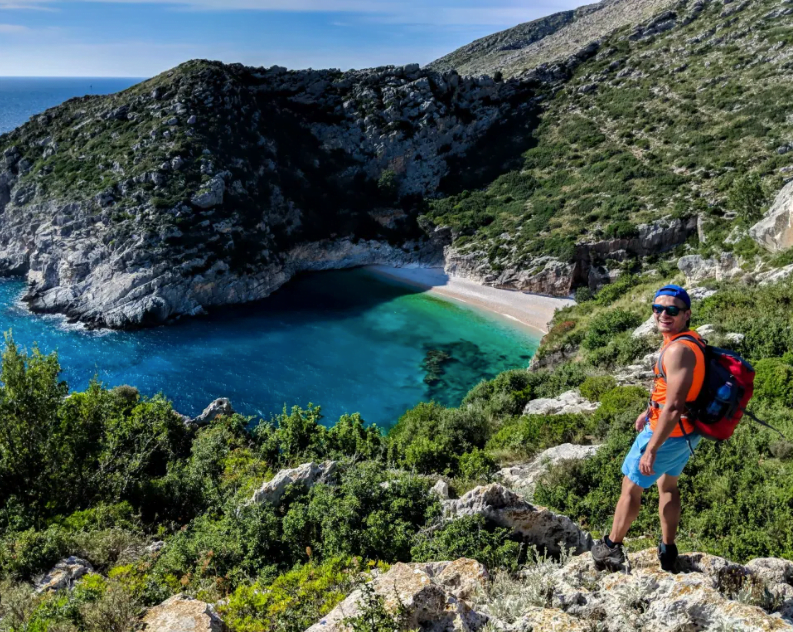 green coast of albania and a man walking in the mountains of it showing a sandy natural beach below and green mountains